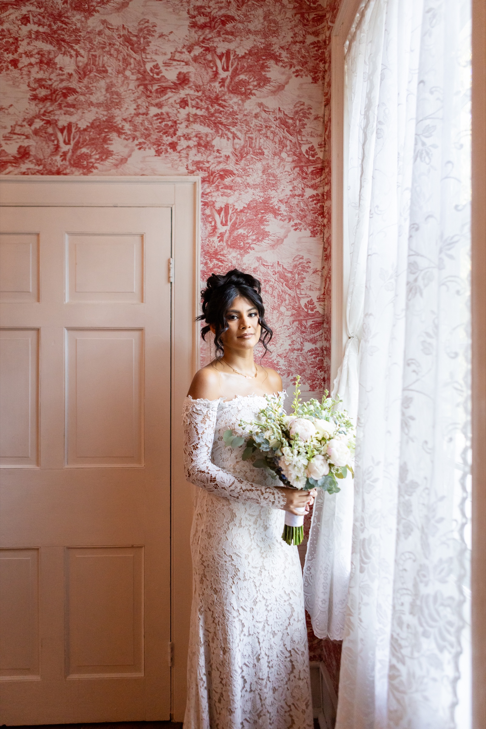 Bride holding bouquet by window with toile wallpaper